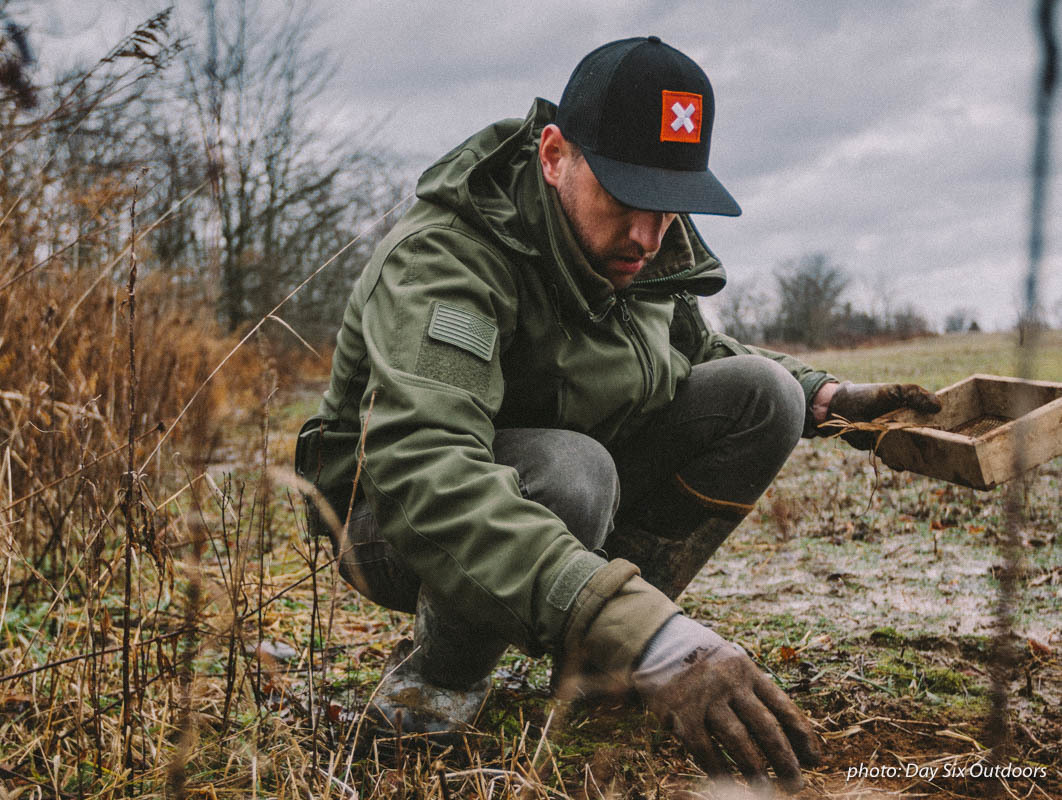 Man in jacket and onX hat camouflaging and setting up animal hunting trap in the woods in the autumn. Man in jacket and onX hat camouflaging and setting up animal hunting trap in the woods in the autumn.