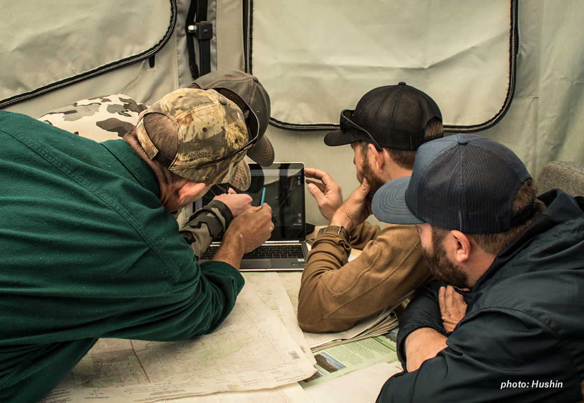 Men gathered in a tent around a computer, e-scouting on onX Hunt Web Map before they go hunting. Men gathered in a tent around a computer, e-scouting on onX Hunt Web Map before they go hunting.
