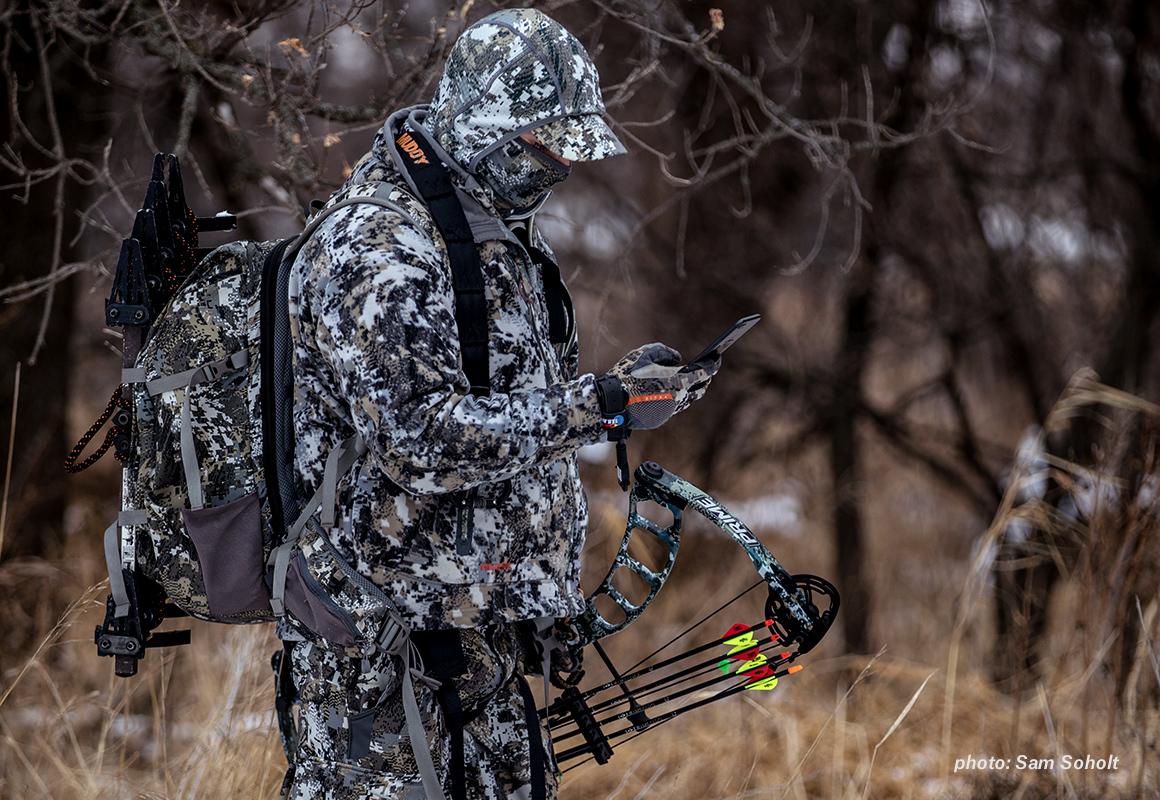 A man in full camouflage and a compound bow checks his onX Hunt App on his mobile phone while hunting. A man in full camouflage and a compound bow checks his onX Hunt App on his mobile phone while hunting.